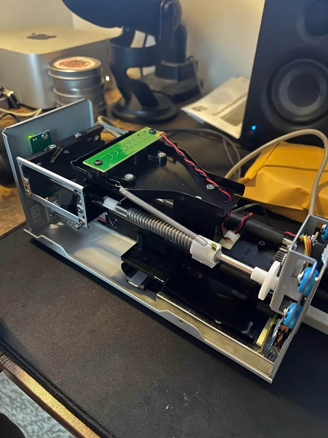 The image shows a disassembled electronic device, possibly a scanner or similar machine, resting on a black desk. The device is primarily silver/grey with a green component on the left side. A black spring is attached to the device, and a white plastic component is visible near the spring. The device is surrounded by v
