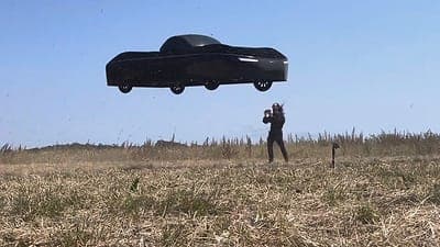 A black car-like vehicle hovers above a dry grassy field under a clear blue sky, while a person holding a controller stands nearby. The scene appears to show a flying-car test.