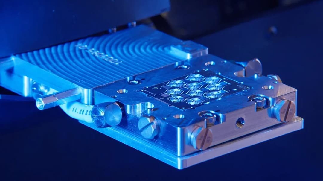 The image shows a close-up view of a metallic device, possibly a part of a machine or tool, illuminated by blue light. The device is rectangular with a slightly curved top and features a grid-like pattern on its surface. It is secured to a dark blue or black surface with several screws and bolts. A flexible hose or tub