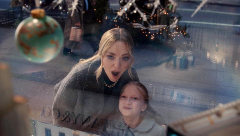 A woman with blonde hair and an open-mouthed expression leans toward a window beside a girl, with a reflected 'TIFFANY & CO.' lettering on the glass, a turquoise ornament, a lit Christmas tree, and a teal gift box in the background.