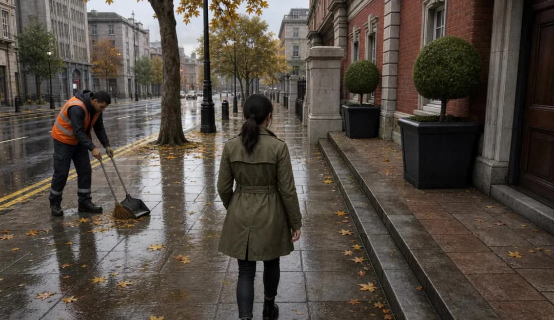The image shows a city street on a rainy day. A man in an orange vest is sweeping the sidewalk, while a woman in a green coat walks away from him. The street is lined with trees and buildings, and the wet pavement reflects the overcast sky. Fallen leaves are scattered on the sidewalk, adding a touch of autumnal color t