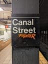 The image shows a black pillar in a subway station, with a sign attached to it that reads "Canal Street Fighter". The sign is primarily white with orange and black text, and it is affixed to the pillar. The background reveals a subway station with a tiled floor and a dark gray or black wall. A portion of a subway platf