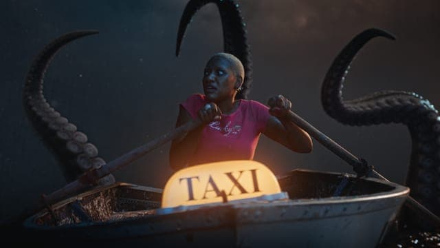 The image shows a woman with short, dark hair, wearing a pink shirt, seated in a small, dark-colored boat. The boat has a yellow taxi sign on its front. The boat is surrounded by three large, dark-colored tentacles, which appear to be octopus tentacles. The background is dark and indistinct, with no clear indication of
