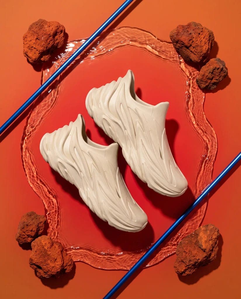 The image presents a pair of beige, high-top sneakers with a unique, wave-like design, positioned on a circular, transparent platform. The platform is surrounded by a ring of orange-red rocks, and two blue lines intersect the platform, creating a geometric pattern. The sneakers appear to be made of a material that rese