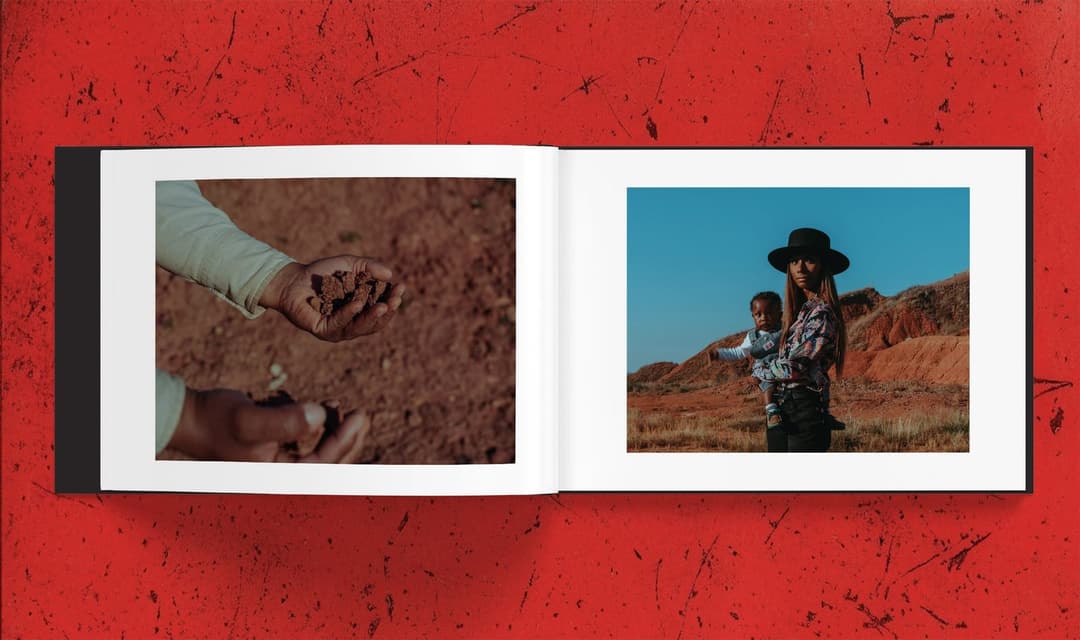 The image shows an open photo album with two pages. The left page features a close-up of a person's hands holding a handful of dark brown soil or dirt. The right page shows a person wearing a black hat and a colorful, patterned shirt, holding a baby in their arms. The background of the right page is a desert landscape