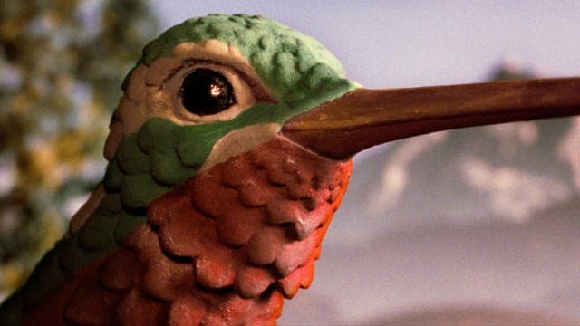 The image shows a close-up view of a bird statue, possibly a hummingbird, with a vibrant mix of red, green, and white colors. The bird's beak is a rich brown color, and its eye is a striking black. The statue is positioned against a blurred background that appears to be a natural setting, possibly a forest or garden. T