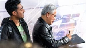 The image shows two men standing in front of a large screen displaying a futuristic design. The man on the left is wearing a black jacket and a green t-shirt, and he is looking towards the screen. The man on the right is wearing a black leather jacket and glasses, and he is holding a stylus and appears to be drawing or