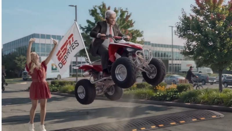 The image shows a man in a suit and tie performing a wheelie on a red and white ATV, with a woman in a red dress standing beside him. The man is holding a white flag with the ESPN Deputies logo. The scene takes place in a parking lot with a building in the background. The ATV is in mid-air, and the woman is standing on