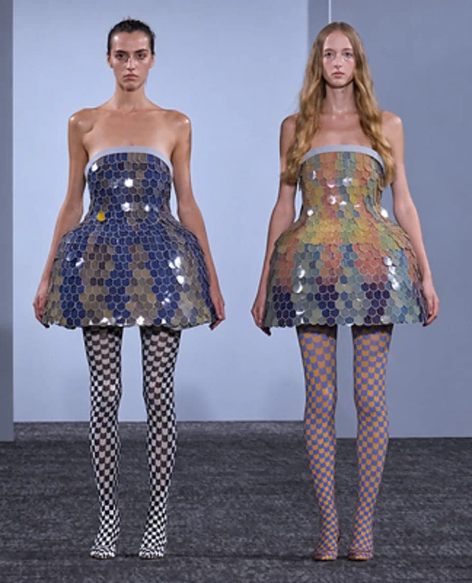The image shows two models standing side by side against a light blue wall. The model on the left is wearing a dress made of small, round, reflective pieces in shades of blue, green, and gold. The model on the right is wearing a dress made of small, round, reflective pieces in shades of orange, yellow, and purple. Both