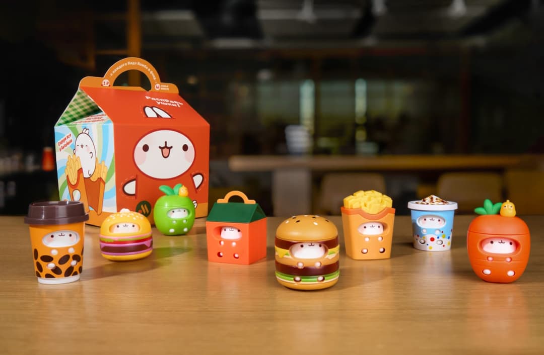 The image shows a collection of colorful toy figurines arranged on a wooden table. The figurines are small and appear to be made of plastic. They are dressed in various outfits, including a green apple, a red and white striped shirt, a red and white striped shirt, a red and white striped shirt, a red and white striped