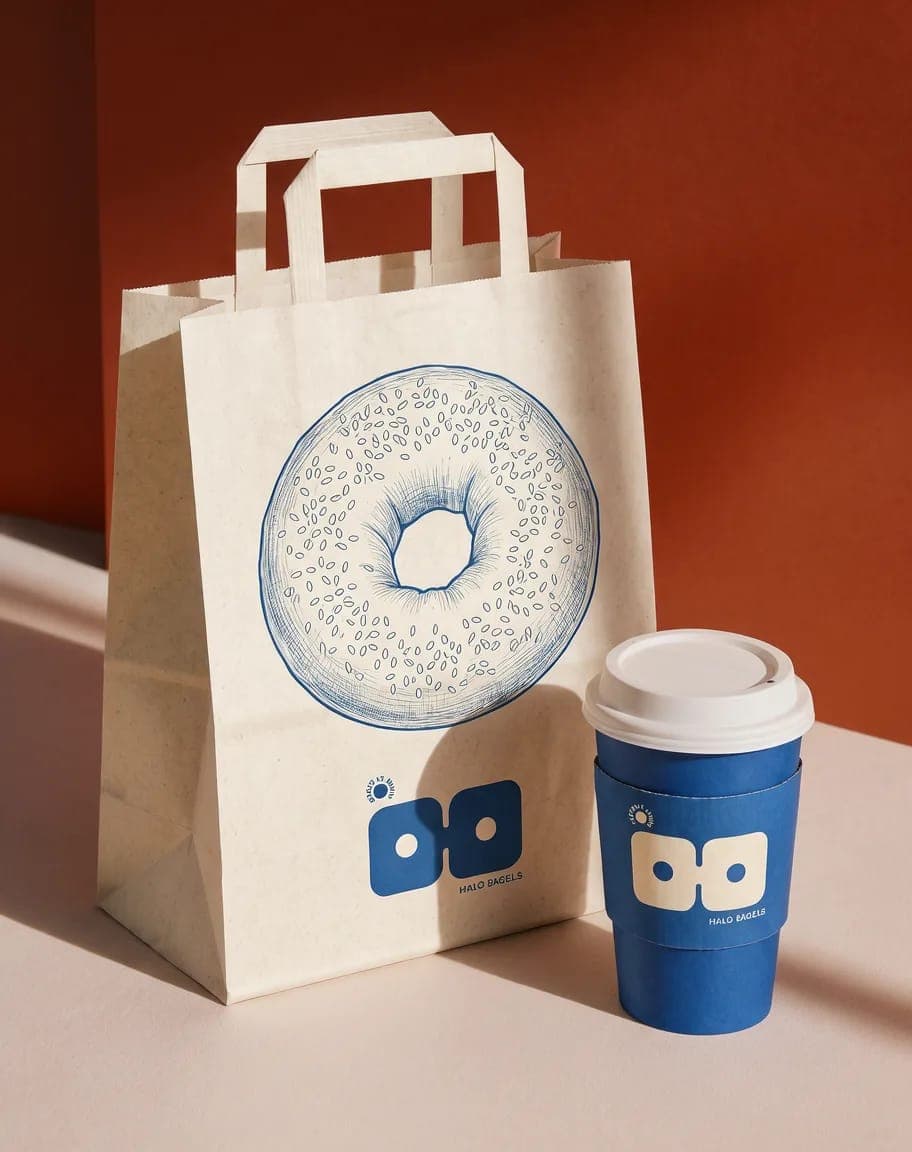 The image presents a white paper bag with a blue illustration of a donut and the text "Halo Bagels" printed on it. The bag is positioned on a light beige or cream-colored surface, with a blue coffee cup featuring the same "Halo Bagels" logo and the text "Halo Bagels" printed on it. The bag and cup are set against a bac