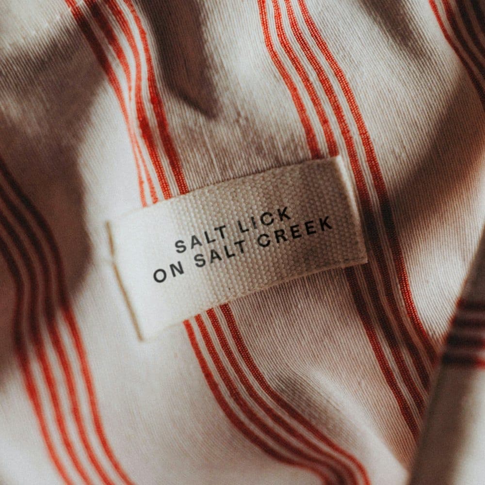 The image shows a close-up view of a fabric with a cream or off-white background and a pattern of alternating red and cream/beige stripes. The fabric has a slightly textured appearance. A white label is attached to the fabric, featuring black text that reads "SALT LICK ON SALT CREEK". The label is positioned in the cen