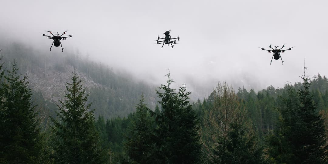 The image shows three black drones soaring in the sky against a backdrop of a foggy mountain. The drones are positioned in a diagonal line, with the first drone on the left, the second in the middle, and the third on the right. Each drone has four propellers and is equipped with a camera. The mountain is covered in a d
