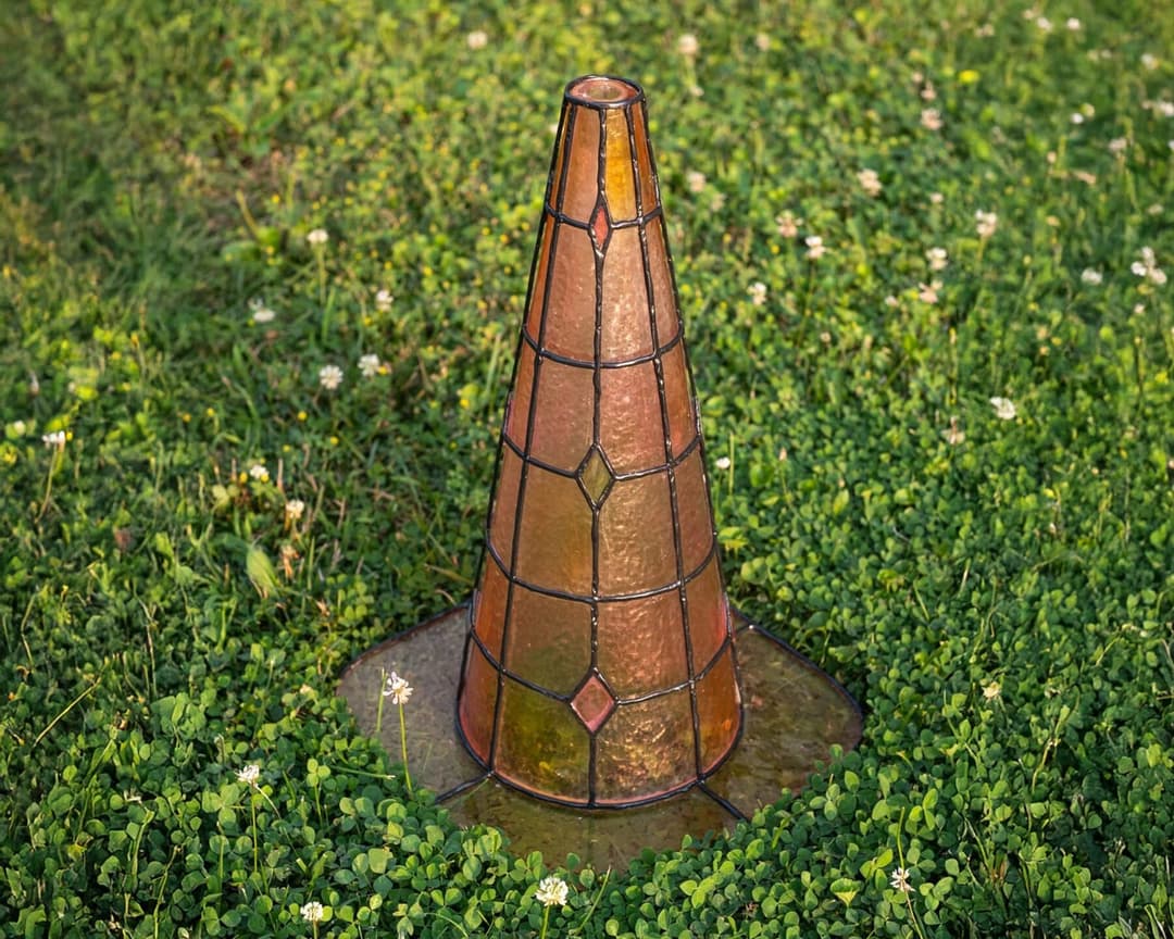 The image shows a copper-colored metal traffic cone with a stained glass design, situated in a grassy area. The cone is slightly tilted to the left and is surrounded by a patch of green grass and small white flowers. The cone is positioned on a small patch of wet ground, possibly a puddle or a small area of water. The