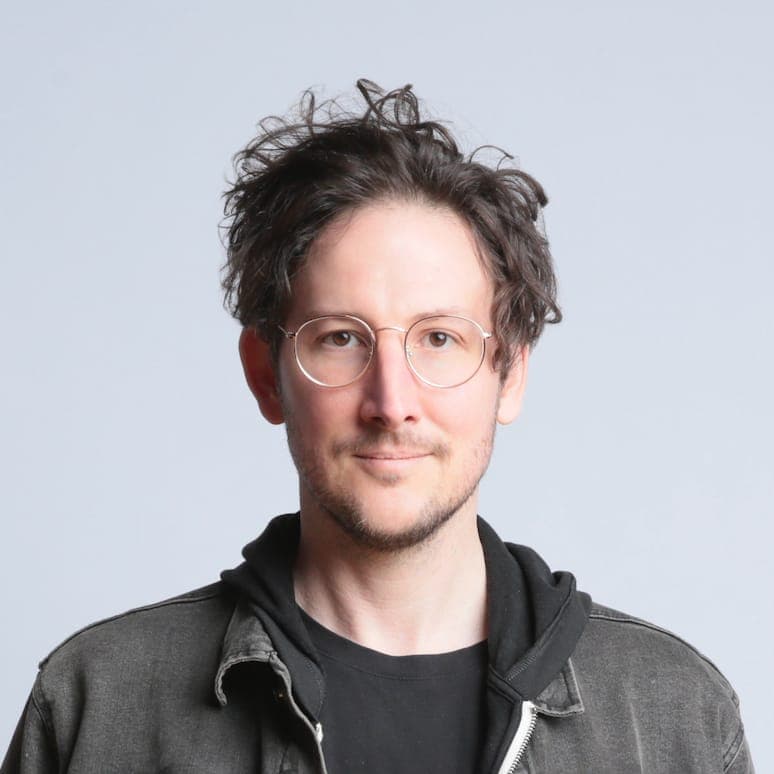 The image shows a man with curly brown hair and a beard, wearing round, wire-rimmed glasses. He is dressed in a black t-shirt and a gray jacket with a black hood. The background is a solid light gray color. The man is looking directly at the camera with a neutral expression.