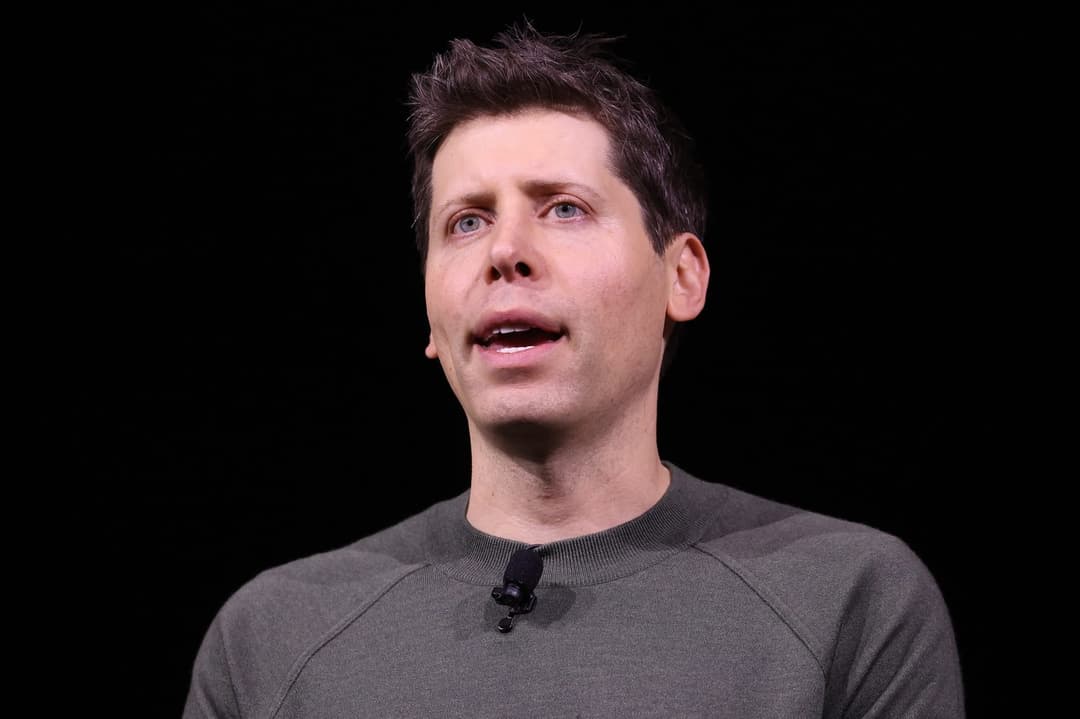 The image shows a man with short, dark brown hair and fair skin, wearing a gray sweater. He is looking upwards and to the left, with his mouth slightly open. A small, black microphone is attached to his shirt near his neck. The background is a solid black, providing a stark contrast to the man's figure.