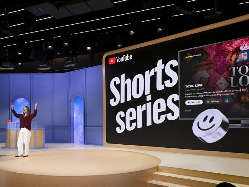 The image shows a woman standing on a stage, with her arms raised in a gesture of excitement. She is wearing a dark red or maroon top and white pants. The stage is set against a backdrop of a blue wall with a circular window. A large screen behind her displays the title "Shorts series" in bold white letters, with the Y