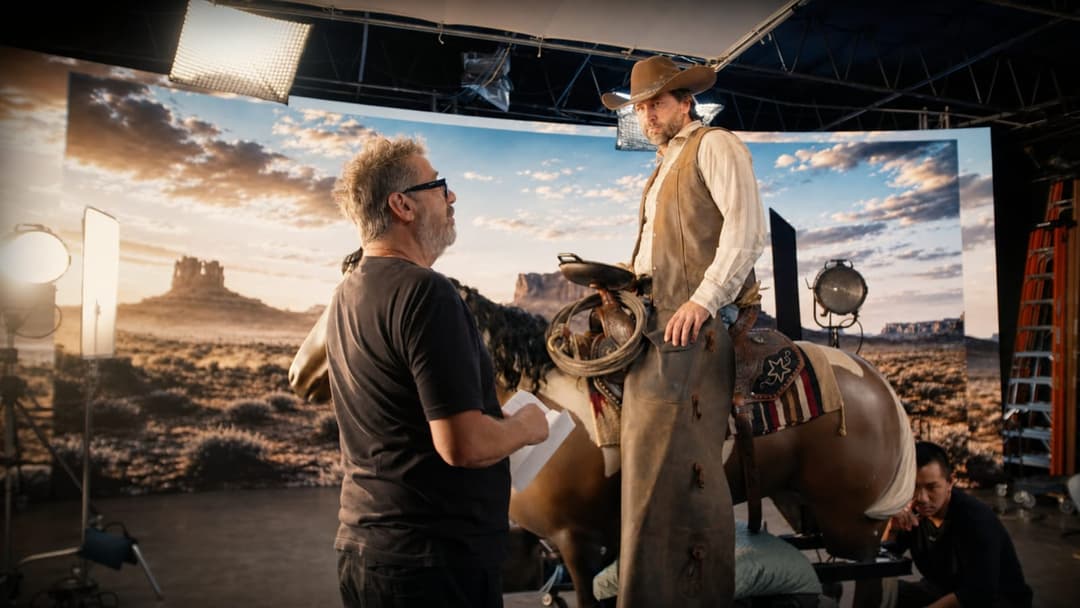 The image shows a man in a cowboy hat and a tan vest standing on a brown horse in a studio setting. The man is wearing a black t-shirt and sunglasses. The horse is adorned with a saddle and bridle. The studio has a large backdrop depicting a desert landscape with a sunset sky. Two men are present in the studio, one sta