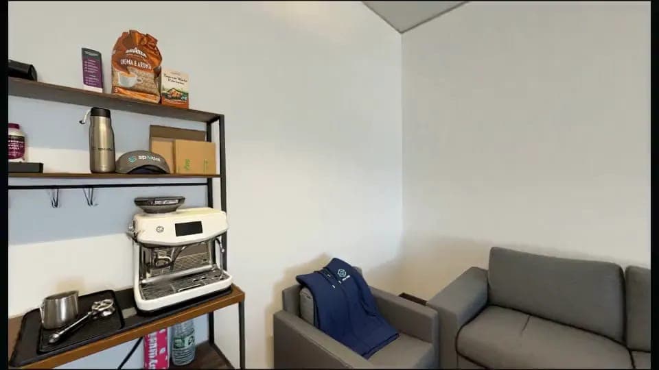 A small office kitchen corner shows a white GE espresso machine on a wooden shelf, with a Lavazza "Crema e Aroma" coffee bag, a stainless steel thermos, and cardboard boxes on the shelves above. A gray sofa and a gray armchair with a blue jacket draped over it sit to the right against a white wall.