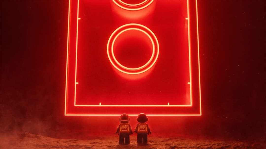 The image depicts two Lego figures standing on a reddish-brown surface, possibly a rocky terrain, with their backs to the viewer. They are wearing white helmets and orange/yellow vests. Above them, a large, rectangular, red LED light displays two circular patterns, one on the left and one on the right. The background i