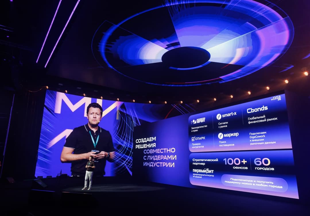 The image captures a man standing on a stage, holding a microphone and gesturing towards a large screen. The stage is bathed in a cool blue light, and the screen displays a vibrant blue and purple abstract design. The man is wearing a black t-shirt and a lanyard with a badge. The screen also features text in Russian, i
