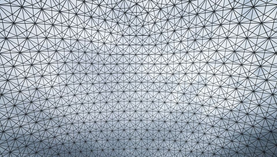 The image presents a geometric pattern composed of black lines forming a network of interconnected triangles. The pattern is symmetrical and creates a sense of depth and dimension. The background is a light blue sky with a few wispy clouds, providing a contrast to the black lines. The image is taken from a low angle, l