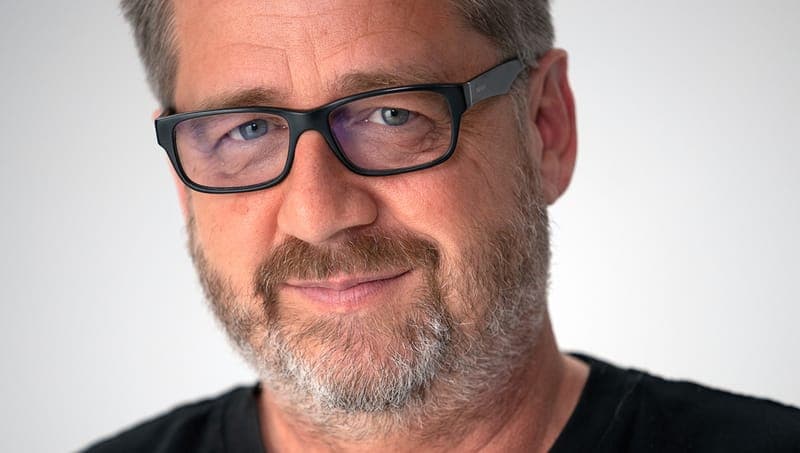 The image shows a man with a warm smile, his eyes looking directly at the camera. He has a light brown or tan complexion and a short, neatly trimmed beard with a mix of gray and white hairs. He is wearing black-framed glasses and a black t-shirt. The background is a plain white, providing a stark contrast to the man's