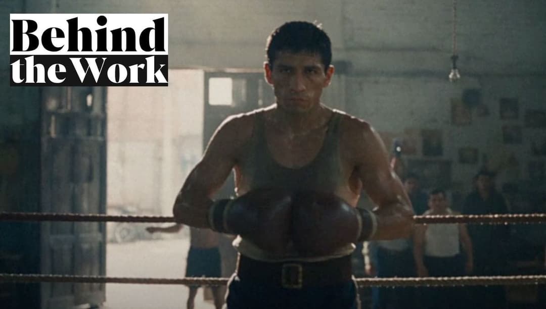 The image shows a man standing in a boxing ring, wearing a gray tank top and boxing gloves. He has dark hair and a serious expression. The boxing ring is made of ropes and has a wooden floor. The background is dark and out of focus, with a few indistinct figures visible. The text "Behind the Work" is superimposed on th