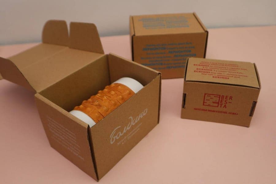 The image shows three cardboard boxes on a pink surface. The largest box is open, revealing a white cylindrical object with an orange textured surface inside. The text on the object is "Baukovo" and "Myr. A.C. Macedonia". The second box is closed and features blue text on its side, which appears to be promotional infor