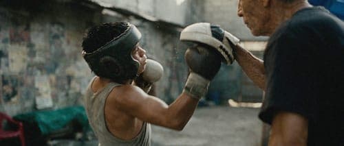 The image shows a young boy wearing a black helmet and a gray tank top, engaged in a boxing practice session. He is wearing boxing gloves and is in the process of throwing a punch towards an older man standing next to him. The older man is wearing a black t-shirt and is also wearing boxing gloves. The scene takes place
