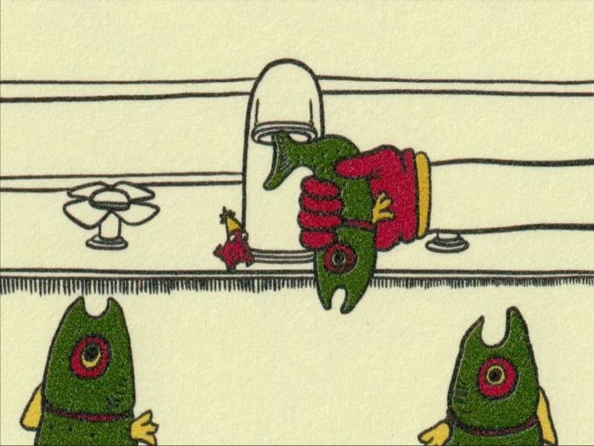 The image depicts a cartoon scene with a hand holding a green fish with a red nose and a yellow fin, positioned on a white sink. The fish is being held by a hand with red gloves. The sink is situated on a white countertop, which is part of a larger white structure. A small, red and white bird figurine is perched on the