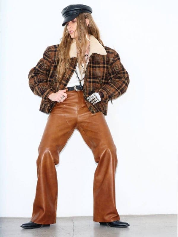 The image shows a person standing against a stark white wall, wearing a black cap, a brown plaid jacket with a white fur collar, and brown leather pants. The person is also wearing a white shirt underneath the jacket and has long, light brown hair. They are wearing a dark-colored belt and have a silver or light-colored