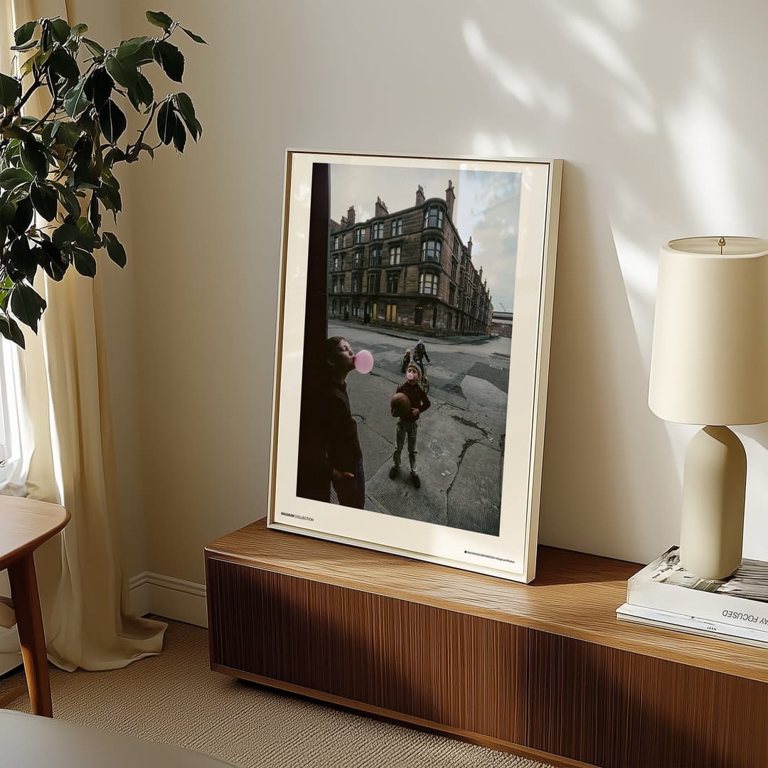 The image shows a living room with a large, rectangular, light-colored picture frame hanging on a white wall. The frame contains a photograph of a child blowing a pink bubblegum bubble on a city street. The photograph is framed in a light beige or cream color. To the right of the frame, a lamp with a cream-colored shad