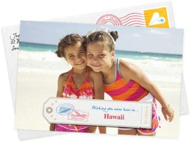 The image shows two young girls standing on a beach, smiling at the camera. The girl on the left is wearing a pink and orange tie-dye dress, while the girl on the right is wearing an orange and pink striped swimsuit. They are holding a white tag with a red stamp that reads "Wishing you were here in Hawaii". The tag als
