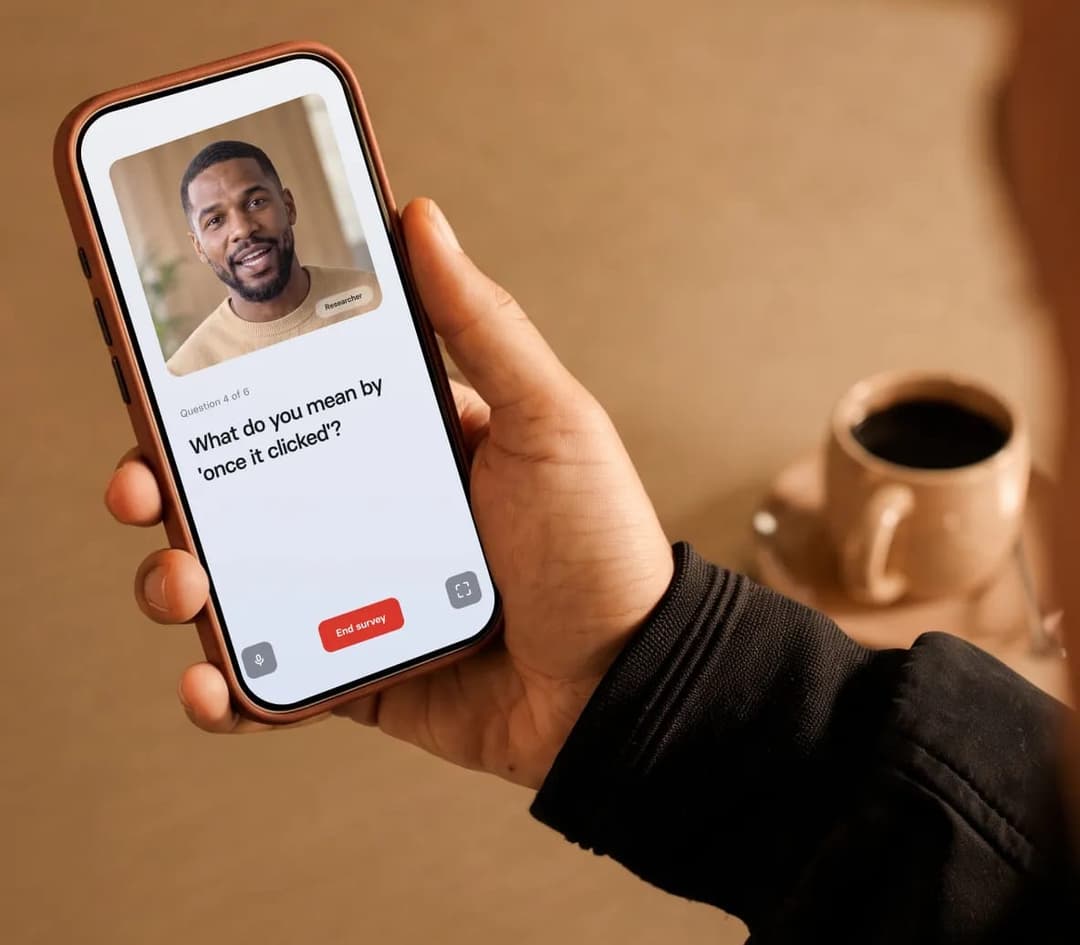 The image shows a person's hand holding a brown smartphone. The phone's screen displays a video call interface with a video call from a man named "Researcher" on the left side. The man is wearing a light-colored shirt and has a beard. The video call interface also includes a "End survey" button. On the right side of th