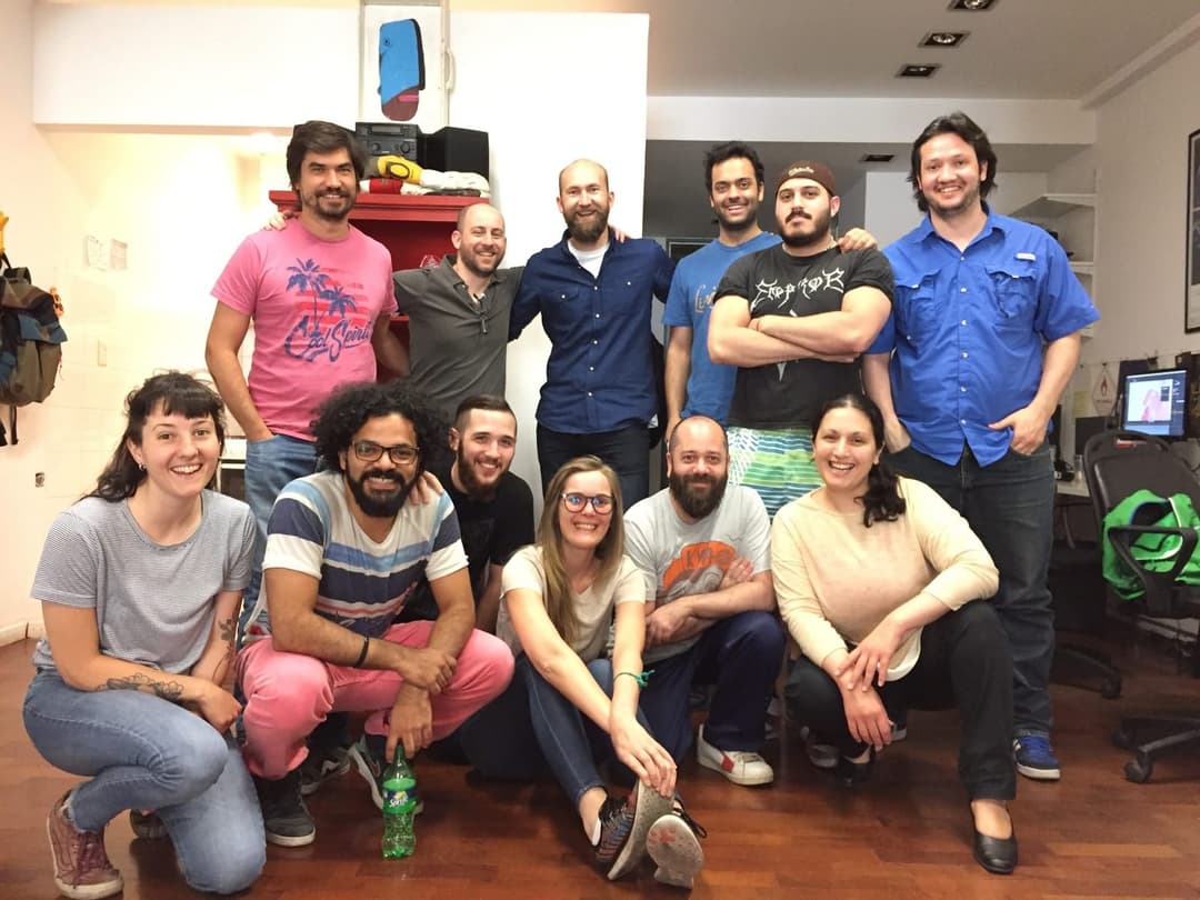 A group of nine individuals, including both men and women, are gathered in a room with a wooden floor and white walls. They are standing in a loose formation, with some individuals standing and others kneeling. The room has a casual, relaxed atmosphere, with a red shelf and a green bottle visible in the background. A t