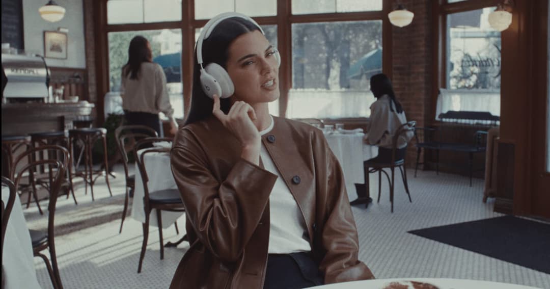 A woman is seated at a table in a restaurant, wearing a brown leather jacket and a white t-shirt. She is wearing white over-ear headphones and is holding them to her ear, suggesting she is listening to music or a conversation. The restaurant has a vintage or retro aesthetic, with wooden tables and chairs, and a brick w