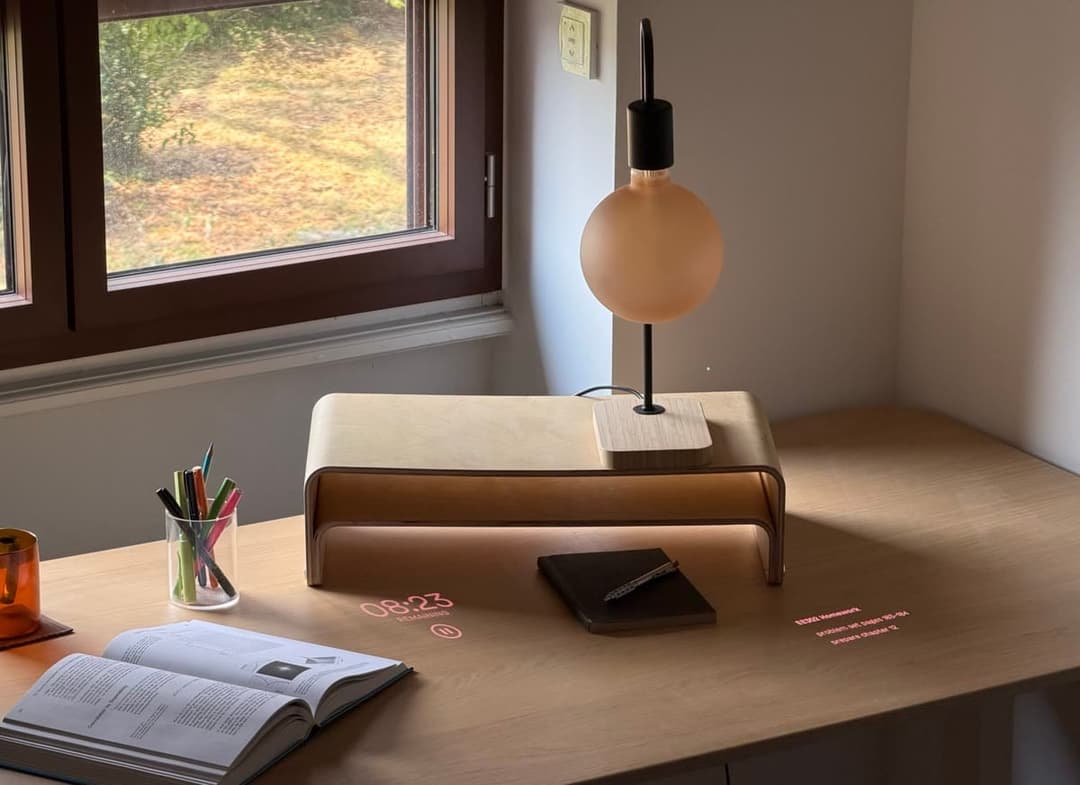 The image depicts a serene workspace setup. A light brown wooden desk is positioned against a white wall, with a window on the left side offering a view of a garden. On the desk, a beige lamp with a black base and a round, beige shade is placed. A black notebook and a pen are also on the desk. An open book with a black
