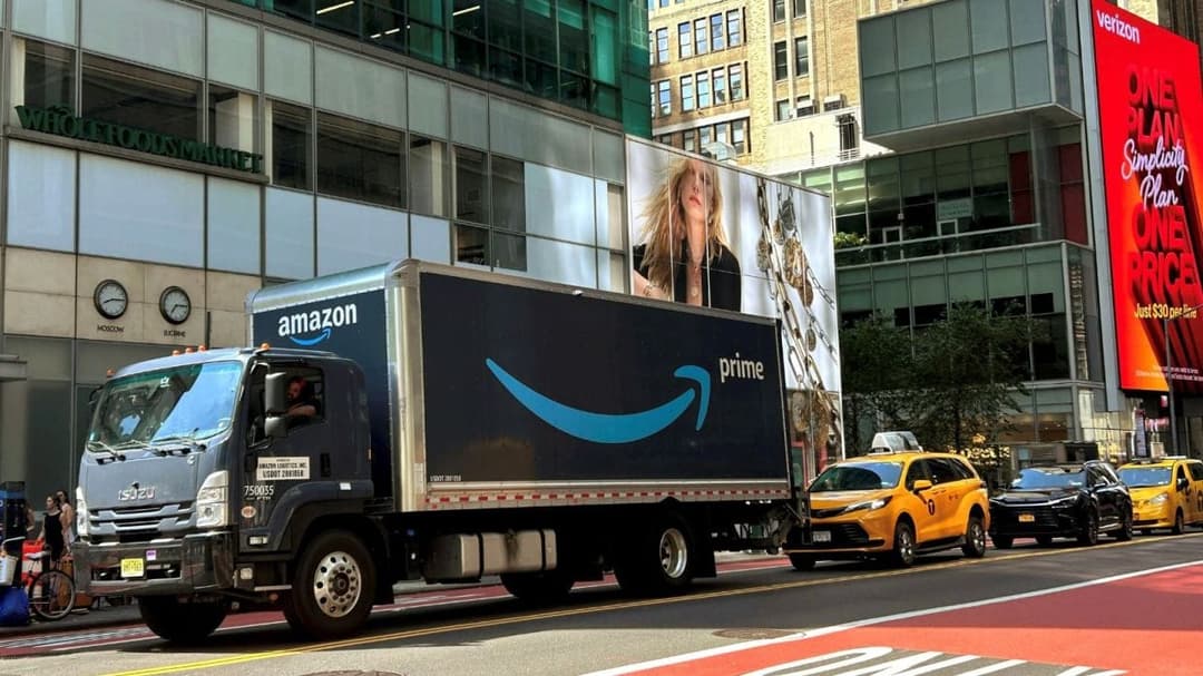 A dark blue Amazon Prime delivery box truck with an Isuzu cab drives down a Manhattan street past yellow taxis, a Whole Foods Market sign, and a red Verizon billboard reading 'One Plan Simplicity Plan One Price, Just $30 per line'.