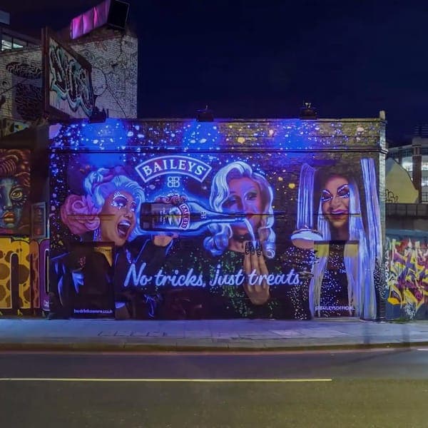 The image depicts a vibrant mural on the side of a building at night. The mural features three women, each with unique hairstyles and makeup, and is illuminated by blue and purple lights. The central figure is holding a bottle of Bailey's No. 1 Irish Whiskey. The women are depicted in a stylized, almost cartoonish mann