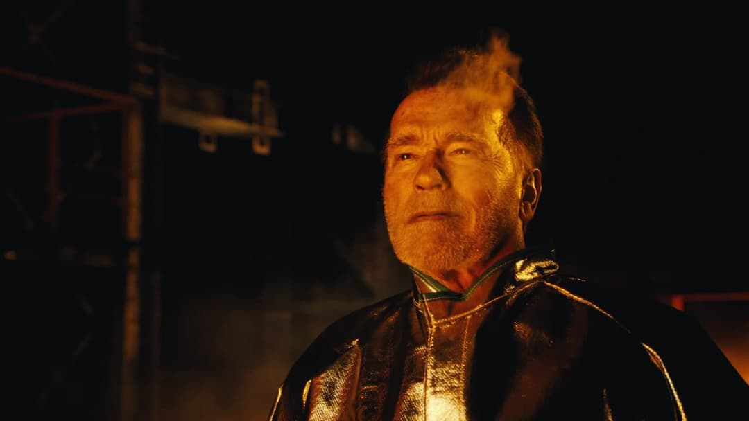 The image shows a man with a beard and mustache, wearing a dark, possibly metallic, jacket with a green collar. He is positioned in the center of the frame, looking off to the side with a serious expression. The background is dark and out of focus, with a hint of a structure or object behind him. The lighting in the im