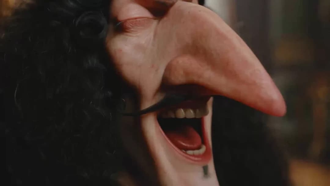 The image shows a close-up of a person's face, with a large nose and a wide, open mouth. The person is wearing a black wig and a mustache, and their eyes are closed. The background is blurred, suggesting a shallow depth of field, and appears to be a dark, possibly indoor setting. The person's skin tone is light.