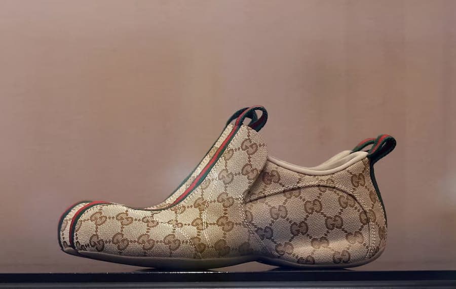 The image shows a pair of beige Gucci shoes with a brown and white patterned canvas, resting on a black surface. The shoes have a slightly curved toe and a pointed toe, and feature a Gucci logo on the side and a Gucci logo on the heel. The shoes are adorned with a red and green Gucci logo on the heel and a red and gree