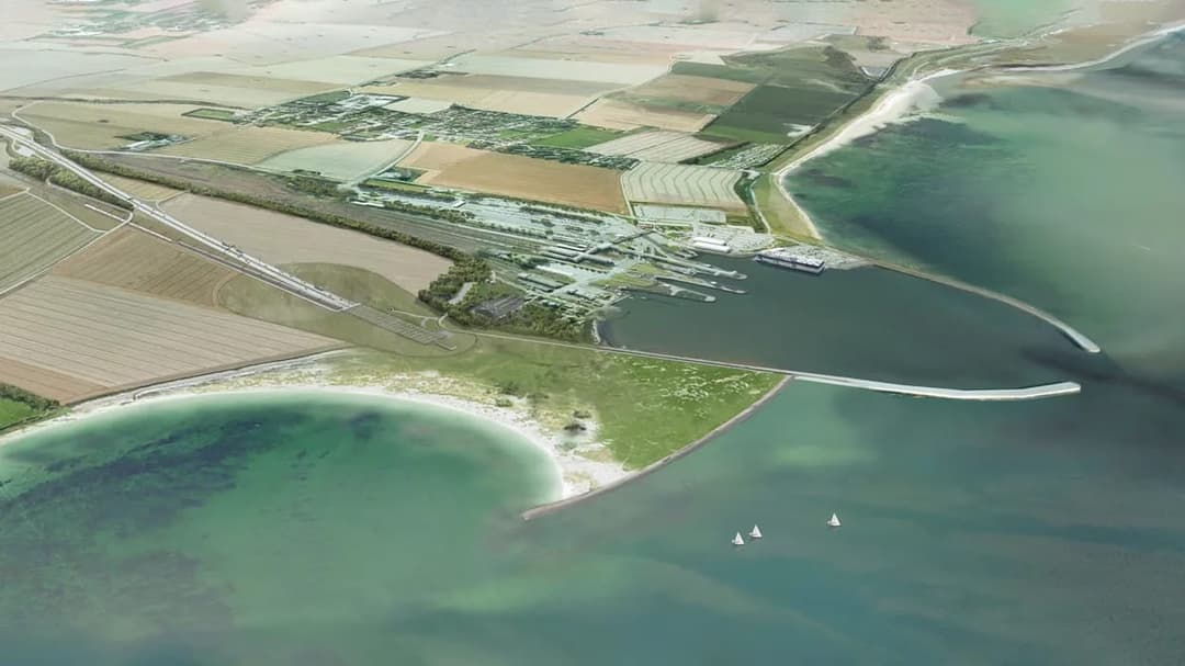The image presents an aerial view of a coastal area, showcasing a mix of green fields, brown agricultural land, and a harbor. A road or highway is visible near the left edge of the image. The harbor is a prominent feature, with several boats visible on the water. A breakwater extends into the water, and a small pier or