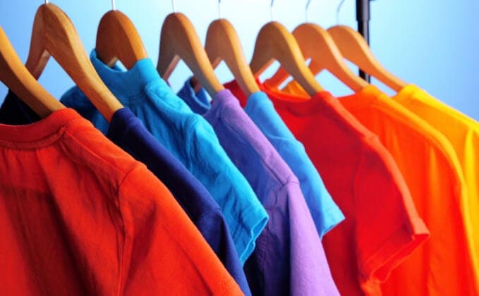 The image shows a collection of colorful t-shirts neatly arranged on wooden hangers against a blue background. The hangers are evenly spaced and support the t-shirts, which are primarily in shades of red, orange, yellow, blue, and purple. The t-shirts appear to be short-sleeved and made of a cotton-like material. The a