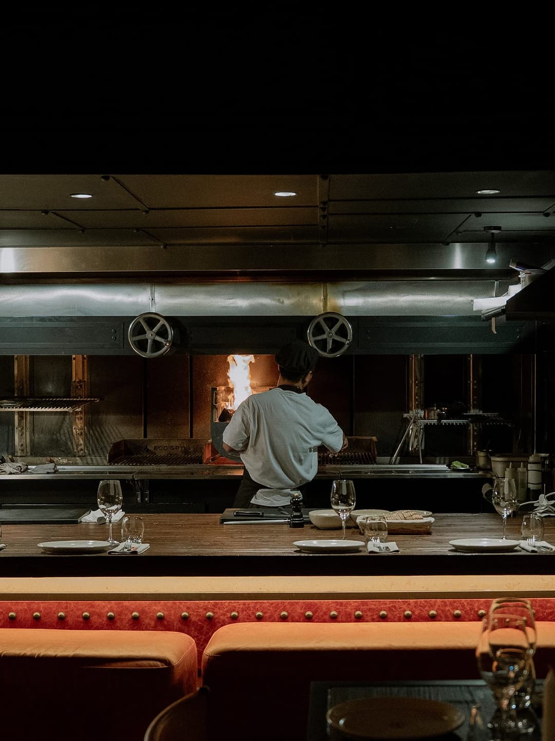 The image shows a chef standing behind a counter in a restaurant kitchen, focused on a large, open fire in the background. The chef is wearing a white shirt and a dark-colored hat. The kitchen features a stainless steel range hood and a large, open fire pit with a fire burning brightly. The counter is made of dark wood
