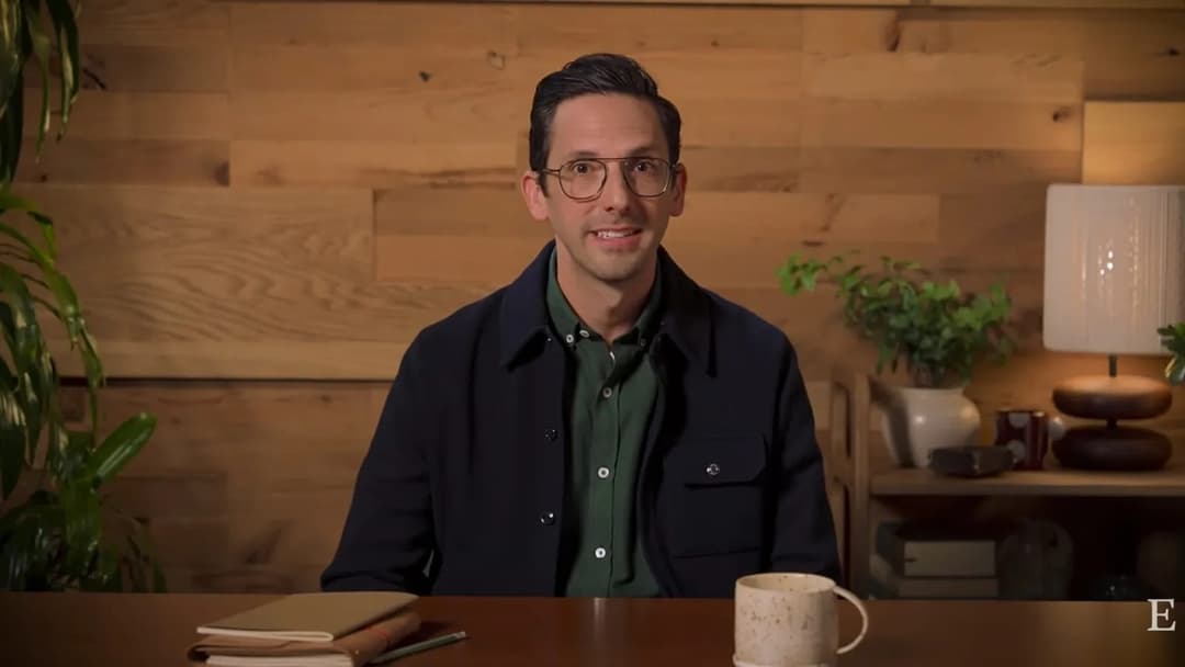 A man with glasses in a navy jacket over a green shirt sits at a wooden table with a notebook, pencil, and speckled mug, in front of a wood-paneled wall and a lamp. An "E" logo appears in the bottom-right corner.
