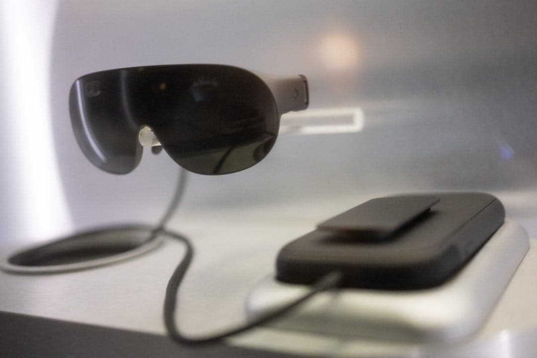 The image shows a pair of black-framed glasses suspended from a wire, which is connected to a black rectangular device, possibly a smartphone or tablet, resting on a white surface. The glasses are positioned slightly off-center, with the wire extending towards the right side of the frame. The background is blurred, dra