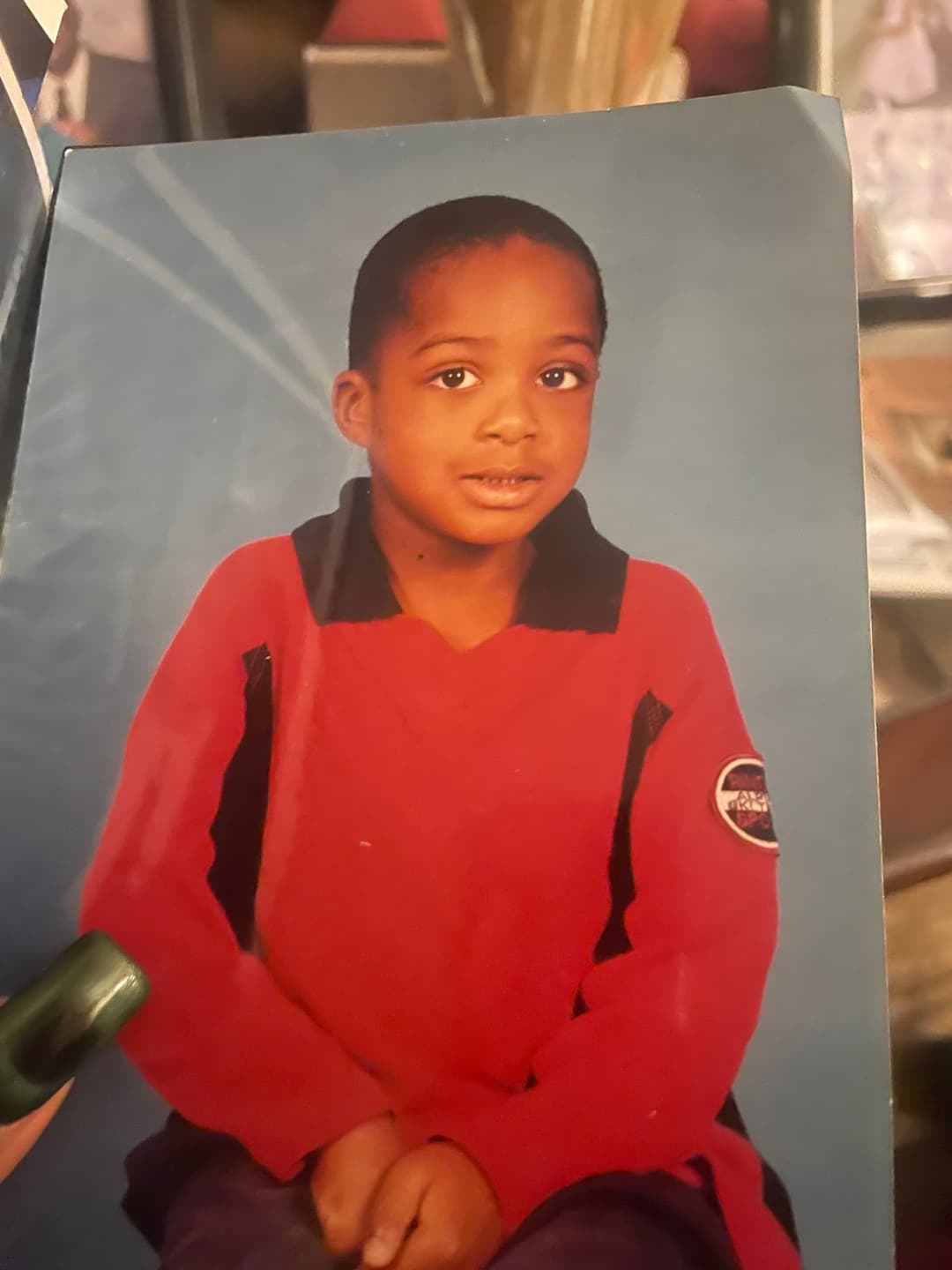 The image shows a young boy with dark brown skin and short, dark hair, wearing a vibrant red shirt with black accents on the collar and sleeves. He is seated and looking directly at the camera with a neutral expression. The boy is positioned in front of a blue background, which appears to be a photo album or similar it