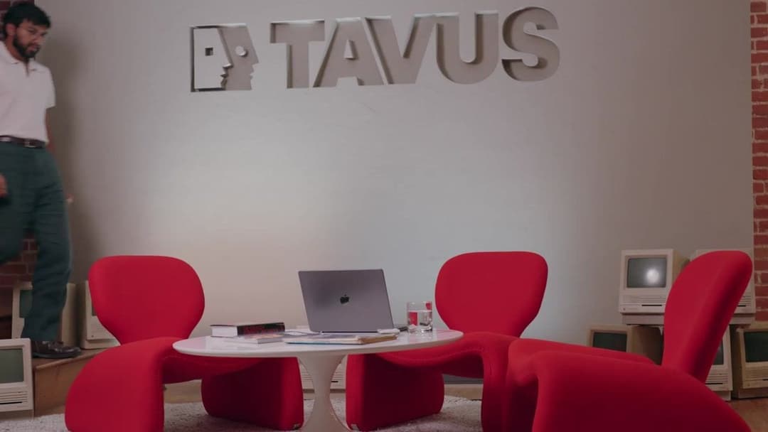 The image shows a modern office space with a white table in the center, featuring a laptop and a glass of water. Three red chairs are arranged around the table, and a white wall with a large, stylized "TAVUS" logo is behind them. To the left of the image, a man in a white shirt and dark green pants is standing. On the